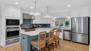 Una cocina moderna con armarios blancos, electrodomésticos de acero inoxidable, una isla central con taburetes y iluminación colgante.