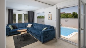 A modern living room with a blue sectional sofa, a small round table, and a sliding glass door leading to a swimming pool. - Casa Crystal Istria (Peroj)