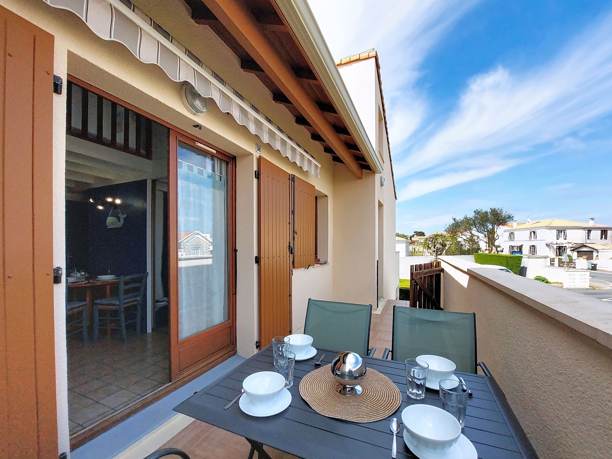 An outdoor dining area with a table set for four, a sliding glass door leading to a dining room, and a view of the street and buildings in the background.