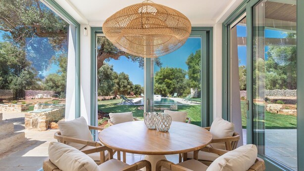 Une table ronde en bois, agrémentée de coussins blancs et surmontée d’un luminaire suspendu en fibres tressées, est entourée de portes vitrées donnant sur un jardin aménagé.