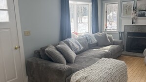 A living room with a grey sofa, a white knitted throw, and a fireplace. - Peaceful 4-bedroom house in charming Castine perfect for relaxing stays (Castine)
