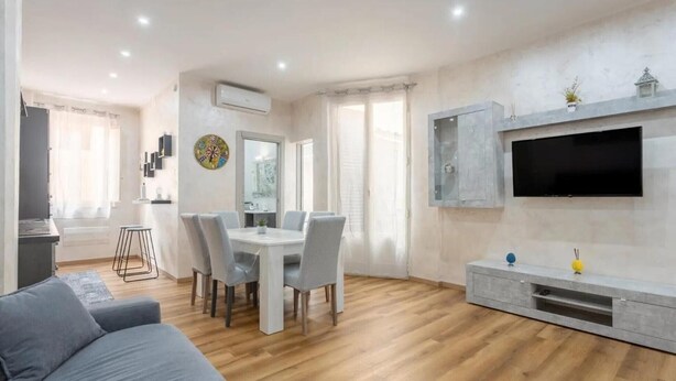 A modern living room with a grey sofa, a dining area with a white table and chairs, a TV mounted on the wall, and a small cabinet below it. - 'Oreto Central Station' Apartment with Balcony, Wi-Fi and Air Conditioning (Palermo)