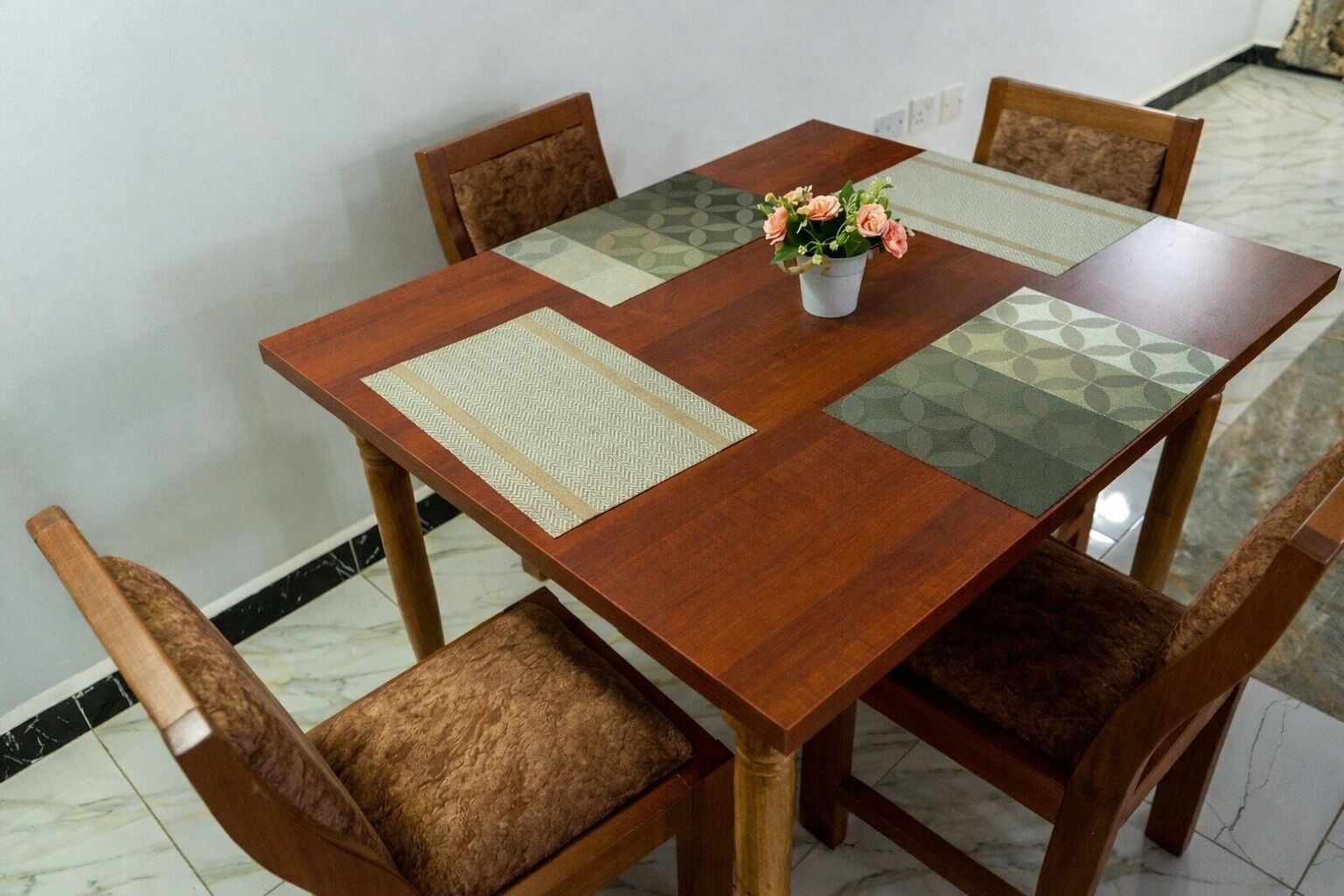 A wooden dining table with four chairs, a vase with flowers, and patterned placemats.