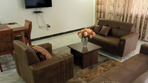 Ein Wohnzimmer mit braunem Sofagarnitur, einem Couchtisch, einem an der Wand montierten Fernseher und einem Essbereich mit Tisch und Stühlen.