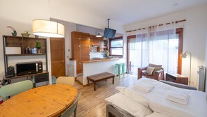 Quarto de hotel compacto com uma mesa de jantar de madeira, uma cama com toalhas cuidadosamente dobradas, uma kitchenette e vista para o exterior através de uma janela.