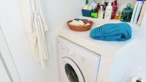 A compact laundry area with a washing machine, a basket of folded clothes, a blue towel, and various toiletries on a countertop. - Hydra studio with patio 250 meters up from port (Ydra)