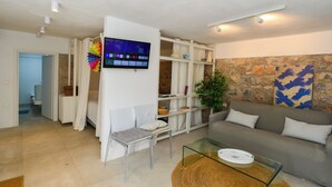 A living room with a sofa, a glass coffee table, a TV mounted on the wall, and a stone wall. - Hydra studio with patio 250 meters up from port (Ydra)