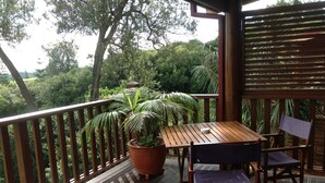 A wooden deck with a table and chairs, surrounded by potted plants and a view of greenery. - Jacaranda Park Holiday Cottages (Norfolk Island)