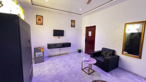 A room with a ceiling fan, a TV mounted on the wall, a sofa, a small round table, and a refrigerator.