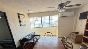 A long wooden dining table with chairs, a ceiling fan, a sofa, a TV, and a window with a view. - Beachfront Apartment : Caleta Beach + Free Parking (Acapulco de Juárez)