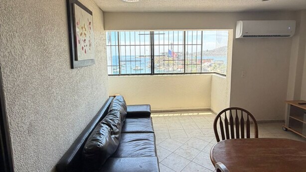 A room with a black leather sofa, a wooden dining table, a chair, a trash bin, and a window with a view of the sea. - Beachfront Apartment : Caleta Beach + Free Parking (Acapulco de Juárez)