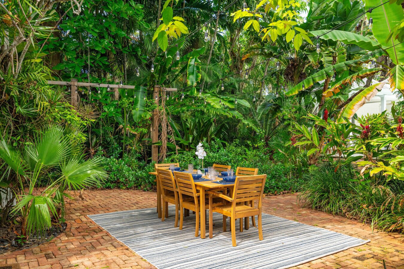 A wooden table set outdoors with chairs, surrounded by lush greenery and a brick pathway.