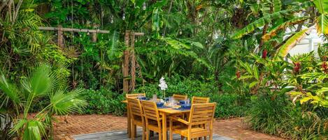 A wooden table set outdoors with chairs, surrounded by lush greenery and a brick pathway.