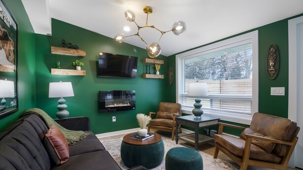 Un salon avec un mur vert, un téléviseur à écran plat, un canapé en cuir marron, deux chaises en bois, une table basse et une lampe.