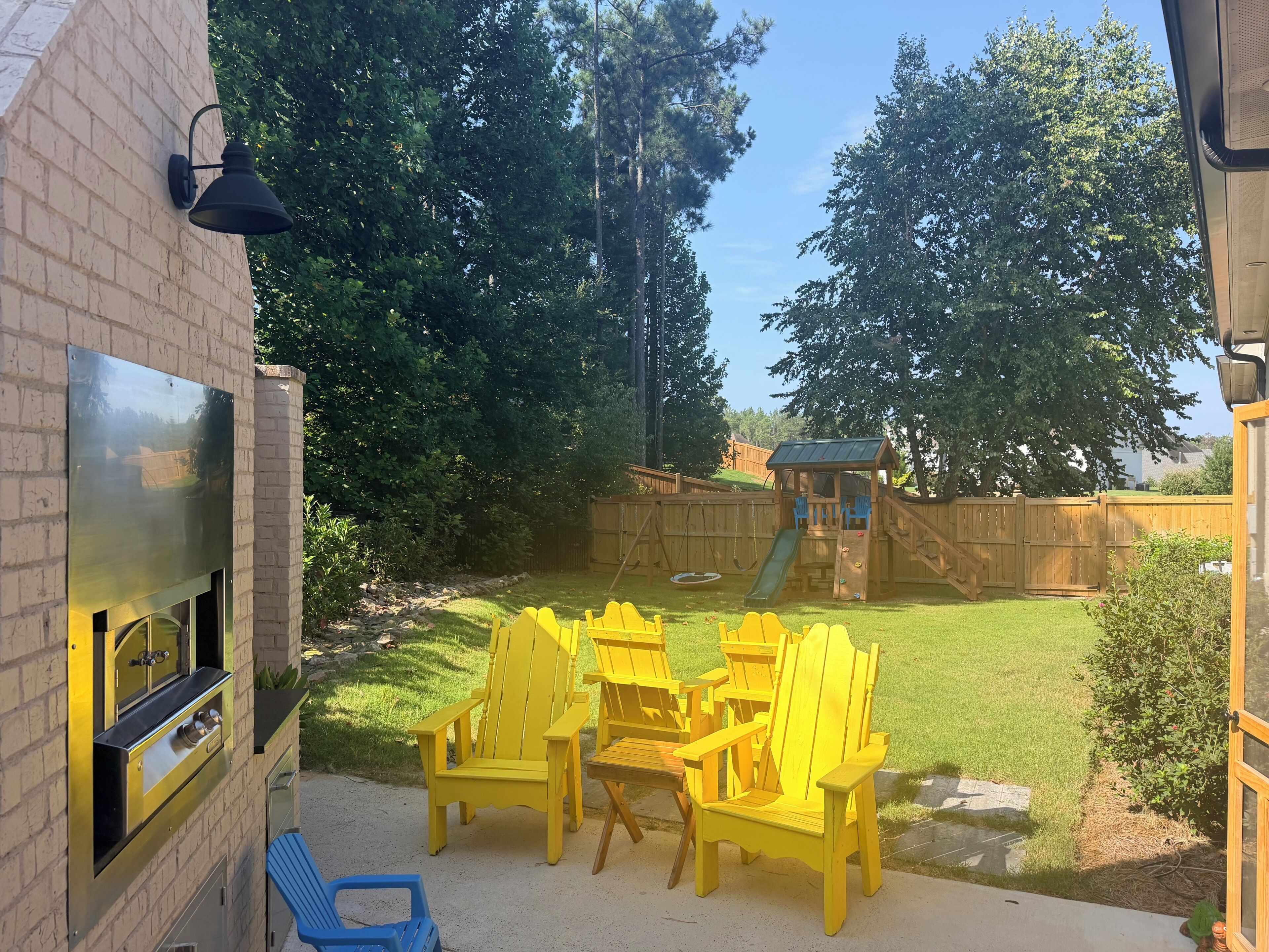 Patio trasero con un parque infantil de madera, una parrilla y sillas amarillas.