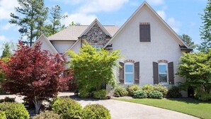 A two-story house with a stone accent on the roof, a driveway, and various shrubs and trees. - Grand Oaks 6BR by Country Club - Sleeps 12 + Crib - Sauna, Pizza Oven, Ole Miss (Oxford)