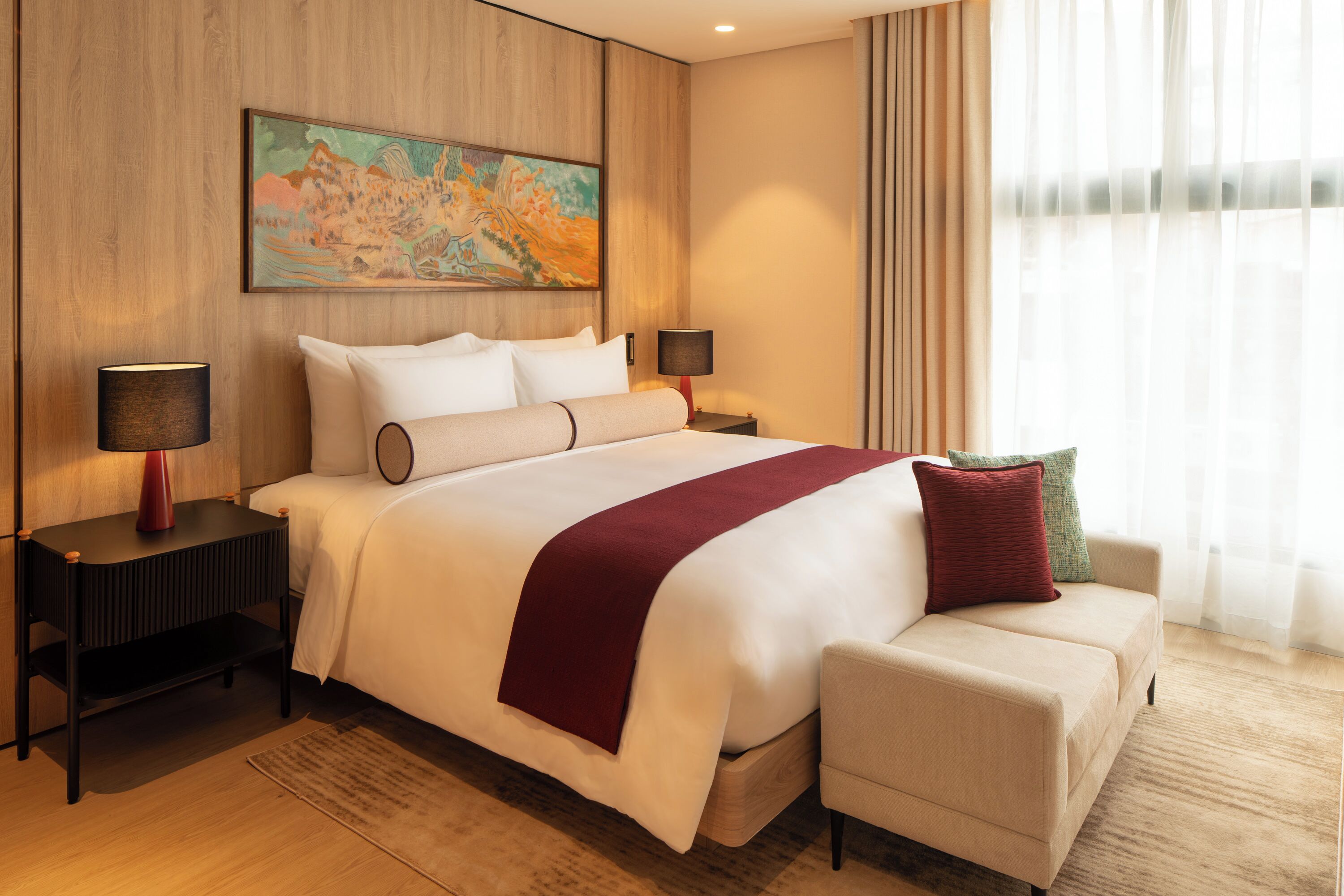 A hotel room with a large bed, two bedside tables, a nightstand, a sofa, a painting on the wall, and a window with curtains.