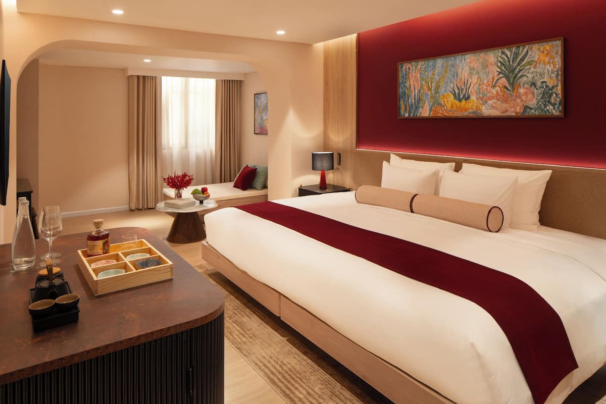 Hotelkamer met een groot bed, een houten nachtkastje met een dienblad met glazen en een fles, een klein rond tafeltje met een bloemenvaas, en een schilderij aan de muur.