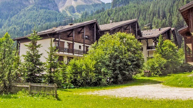 Un edificio de estilo chalete tradicional con fachada de madera, frente a imponentes montañas verdes y un cielo despejado.