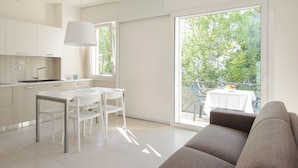 A modern kitchen with white cabinetry, a dining area with a white table and chairs, and a balcony with a table and chairs. - Lignano Apartment With Balcony (Lignano Sabbiadoro)