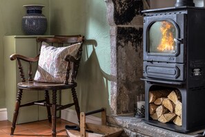 A wooden chair with a patterned cushion, a black stove with visible flames, and a stack of firewood. - Miners Cottage (2) - sleeps 2 guests  in 1 bedroom (Bakewell)