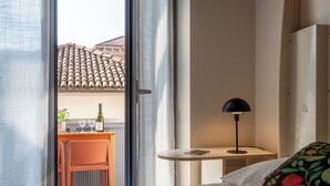 A hotel room with a bed, a desk with a chair, a lamp, and a view of a building with a tiled roof. - Domus Talenti by Interhome (Alba)