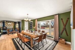 A dining area with a wooden table set for six, surrounded by chairs, with a view of a street through large windows.