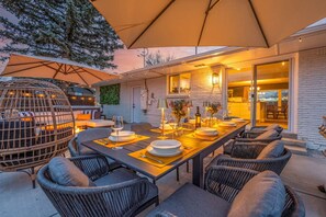 Uma área de refeições ao ar livre com uma mesa comprida, cadeiras e churrasqueira.