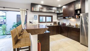 A modern kitchen with dark wood cabinets, a stainless steel refrigerator, and a dining area with wicker furniture. - Huge Private Pool Villa Atala Canggu Seminyak 4BR (Kerobokan)