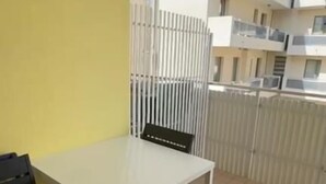 Een balkon met een witte tafel en stoelen, een gele muur en uitzicht op aangrenzende gebouwen.