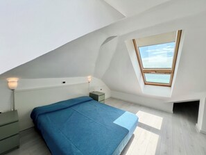 Ein Schlafzimmer mit schräg verlaufender Decke, einem Oberlicht, einem Bett mit blauer Bettdecke, Nachttischen und einem Nachtschränkchen.