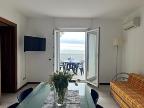 A room with a dining table, chairs, a sofa, a TV, and an open door leading to a balcony with a view. - Lignano Beachfront Apartment (Lignano Sabbiadoro)