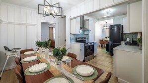 Een eetgedeelte met een houten tafel voor zes personen, een hoog plafond met een geometrische lamp, en een keuken op de achtergrond.