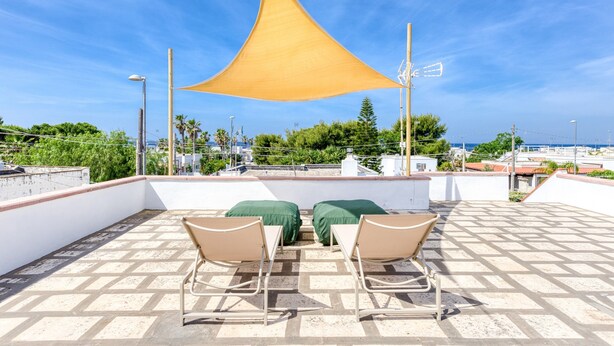 A rooftop terrace with two chairs, a yellow shade, and a view of the sea and buildings.