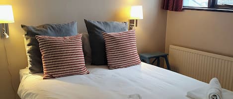 Una cama bien tendida con sábanas blancas, dos almohadas estampadas y dos toallas enrolladas.