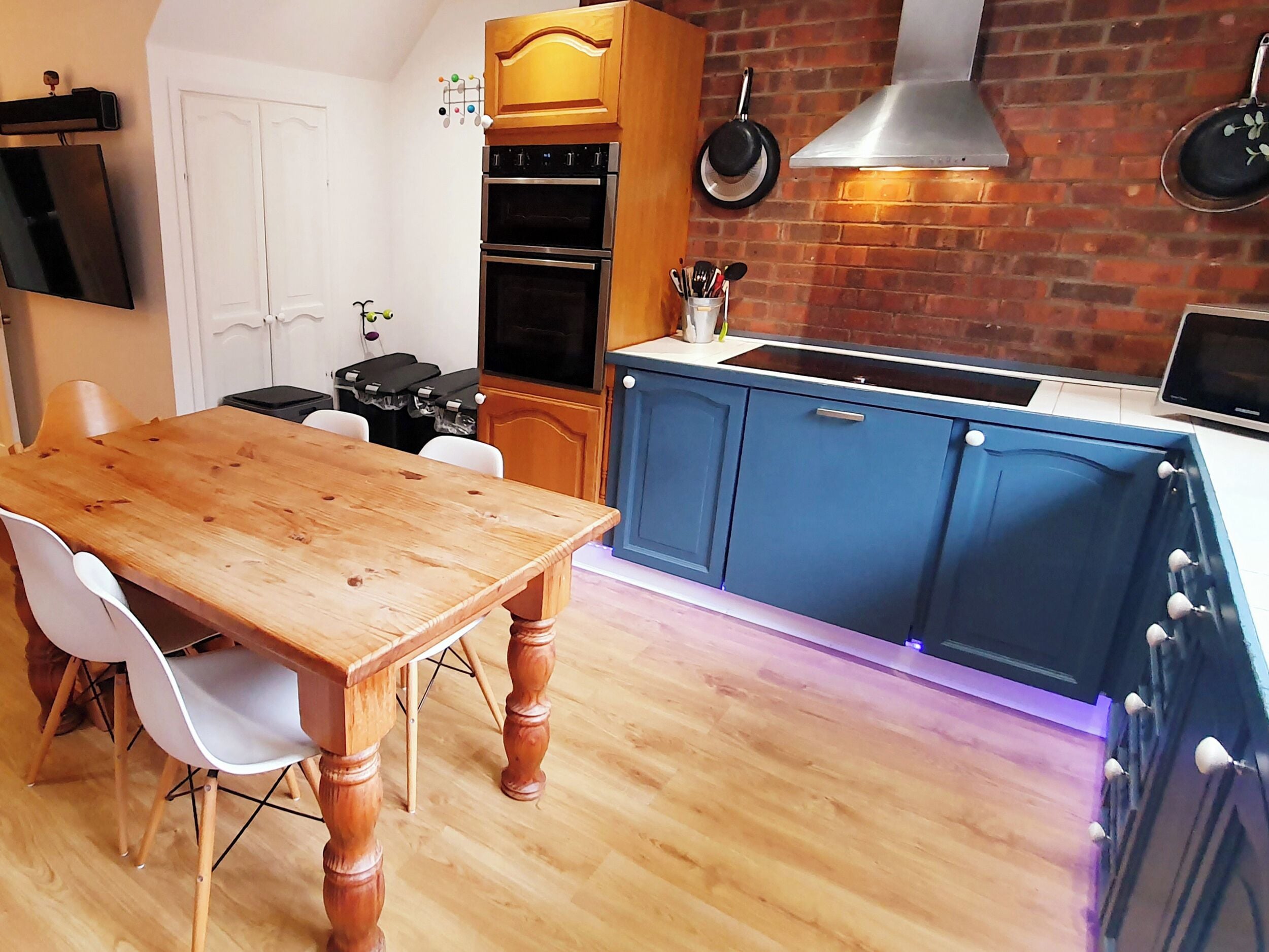 A kitchen with wooden cabinets, a dining table, chairs, a TV, a stove, and a brick wall.