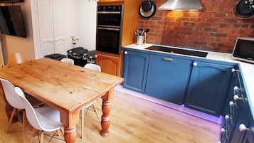 A kitchen with wooden cabinets, a dining table, chairs, a TV, a stove, and a brick wall.