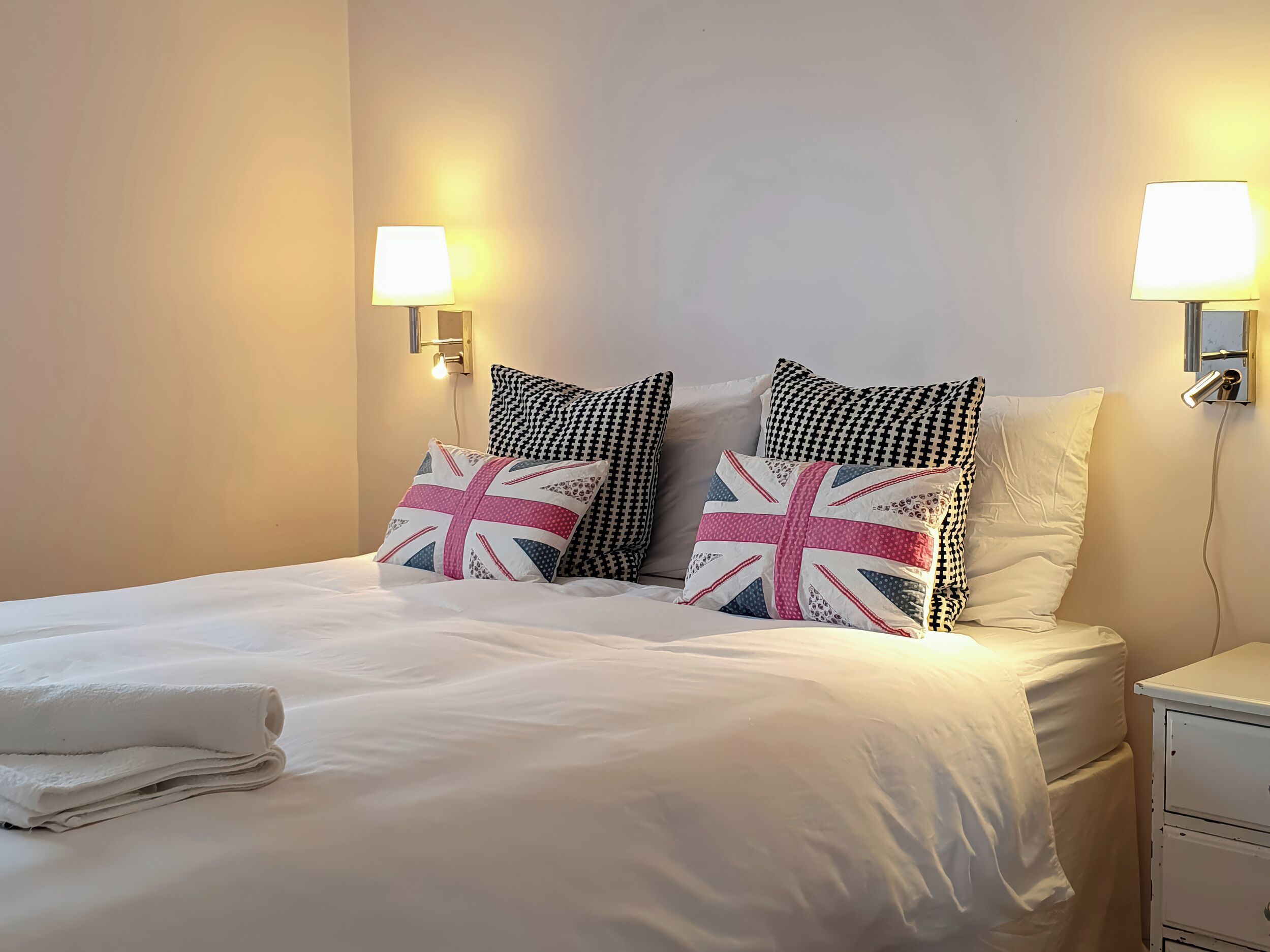 Un lit bien fait, avec du linge de lit blanc et deux oreillers ornés d’un motif Union Jack, une table de chevet avec une lampe et un luminaire fixé au mur.