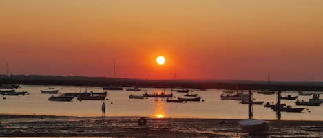 A sunset over a calm body of water with boats and a person standing on the shore.