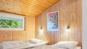 Quarto com paredes de madeira, duas camas com roupa de cama branca, um quadro na parede e uma janela com vista para o verde.