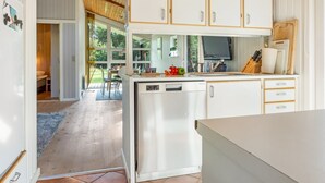 Cozinha compacta com armários brancos, uma pequena geladeira e uma área de refeições visível por uma porta aberta.