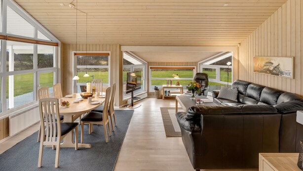 Een ruime woonkamer met een eettafel, stoelen, een leren bank en grote ramen met uitzicht op de omgeving.