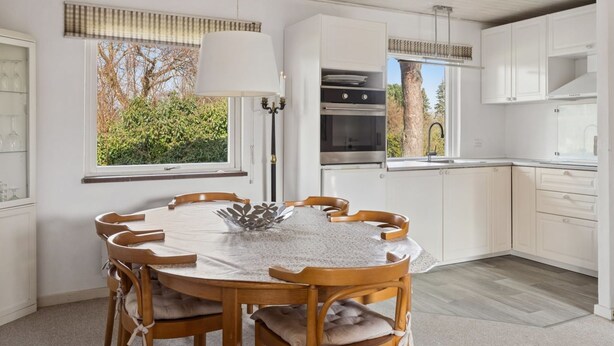 A modern kitchen with white cabinetry, a dining table with chairs, a pendant light, and a view of greenery outside.