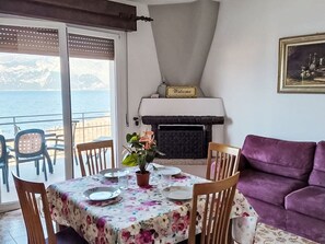 Un coin repas avec une table dressée pour quatre personnes, un canapé violet, une cheminée et une vue sur la mer.