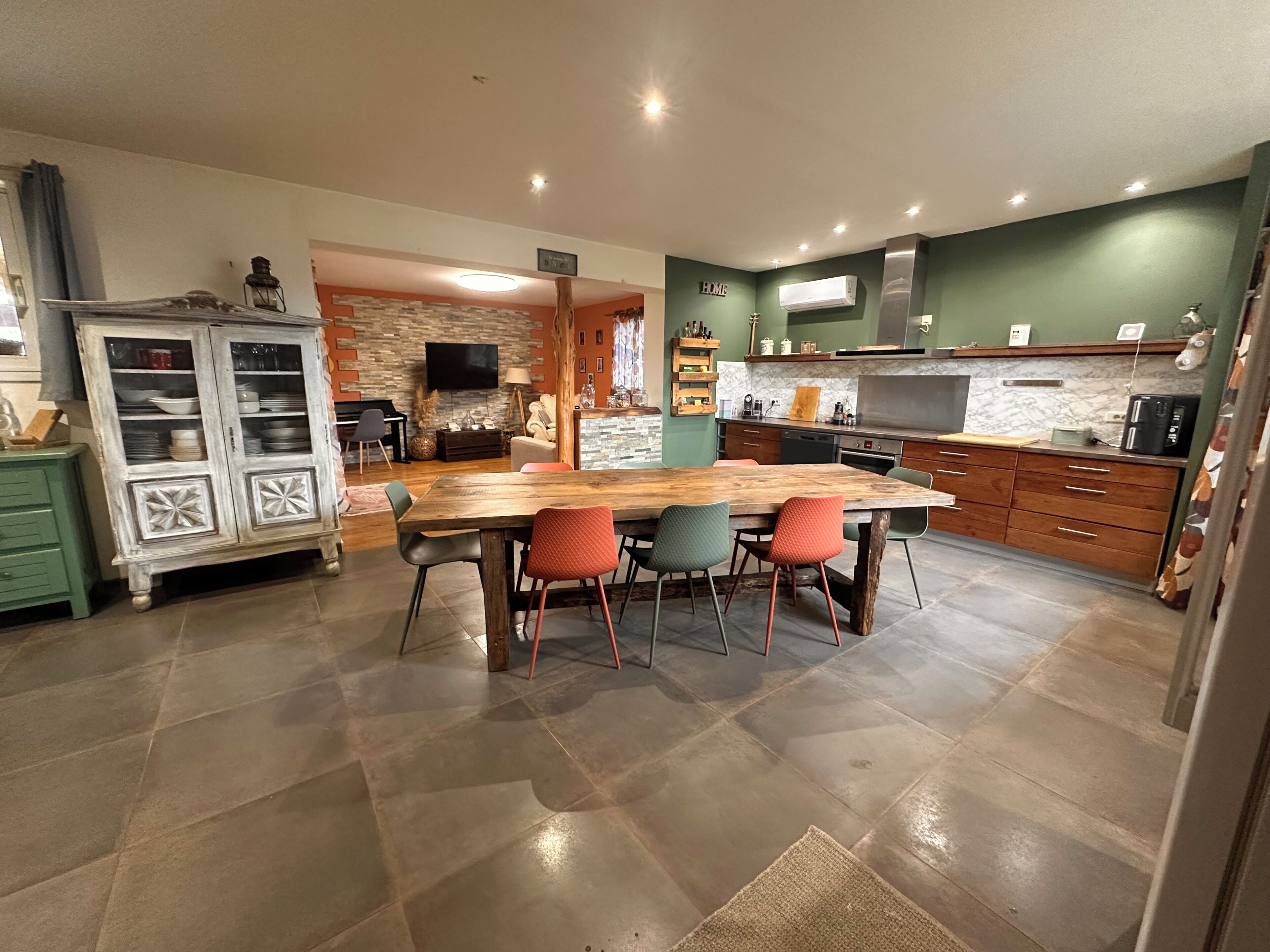 A modern kitchen with a large wooden table, four chairs, a built-in oven, and a TV mounted on the wall.