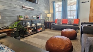 A living room with a sofa, ottomans, a coffee table, a bookshelf, and a window with curtains.