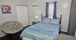 Une chambre à coucher comprenant un lit, une chaise, une table de chevet avec une lampe, une fenêtre avec des rideaux et un climatiseur fixé au mur.