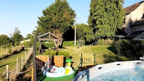 Une piscine avec un escalier, une structure de jeu en bois et une cour gazonnée avec des arbres.