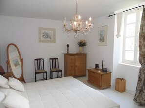 Un dormitorio con cama, dos sillas, cómoda, espejo, candelabro y obras de arte en las paredes.