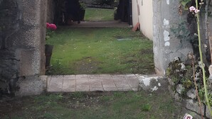Una estructura de piedra en arco con un área verde al frente, un muro de ladrillo y una planta con flores rosadas.